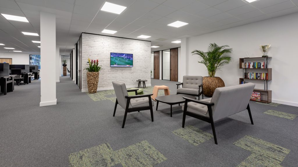 centre of an office. desks and computers on the left of the office and a breakout space in the middle, with grey chairs and furniture, and a TV.
