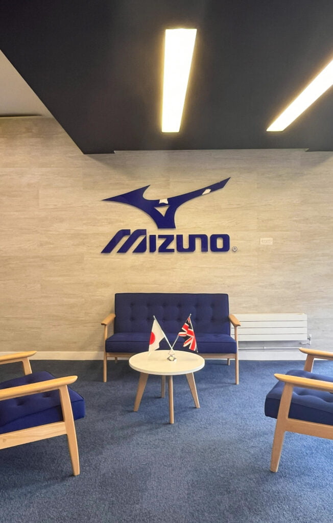 mizuno reception area with blue sign, blue chairs, blue carpet and white coffee table with UK and Japanese flag. 