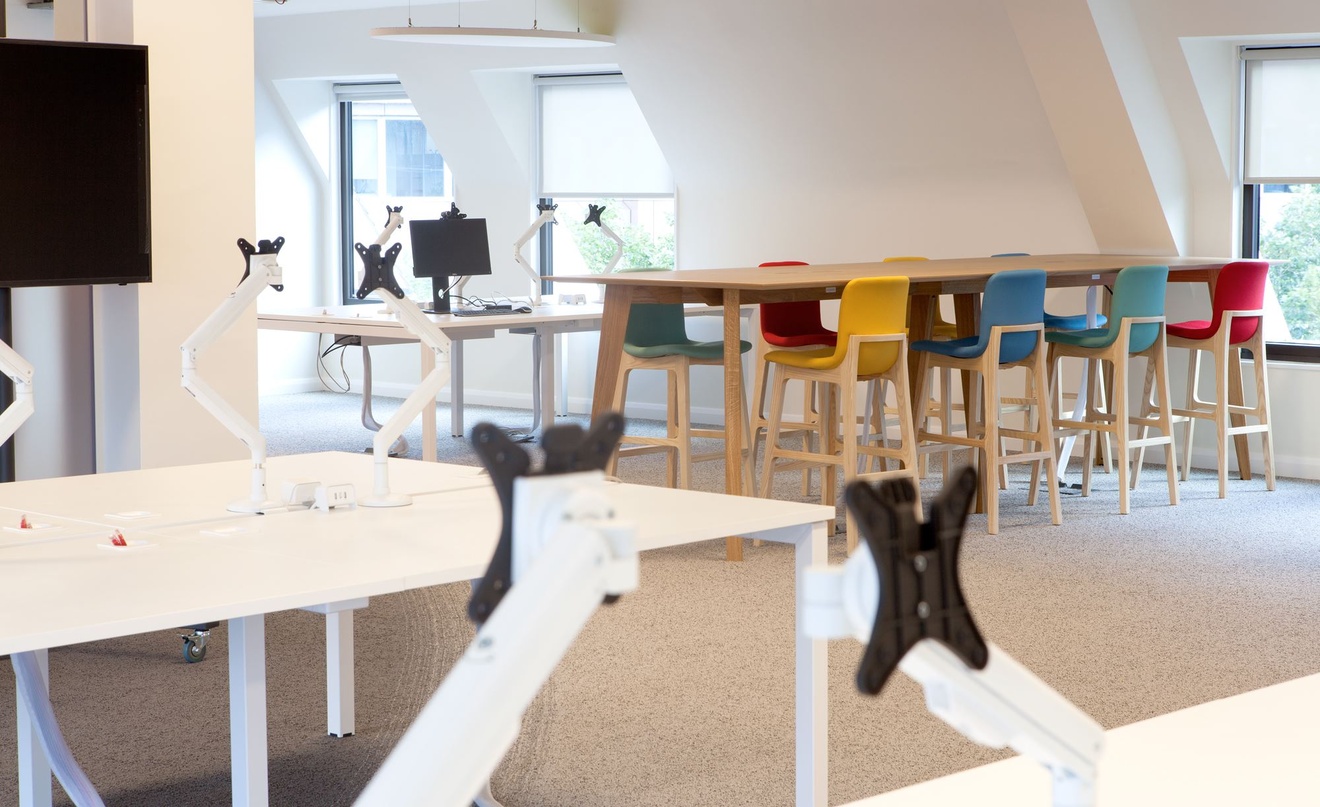 Office desk with monitor brackets on top and a table with stools in the background.