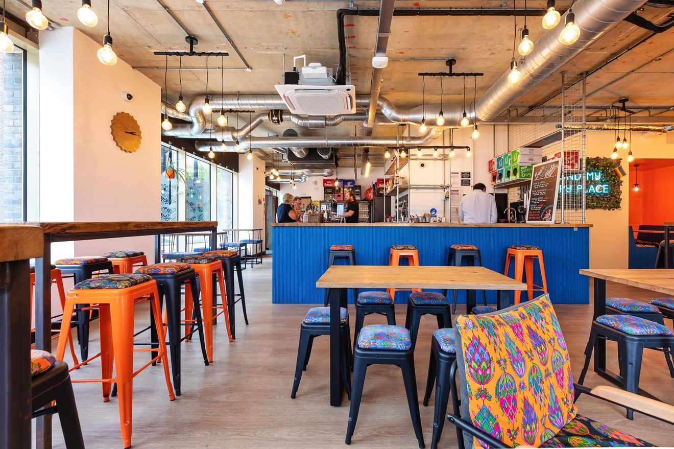 A restaurant with two people stood ordering a the bar and empty tables and chairs surrounding them.