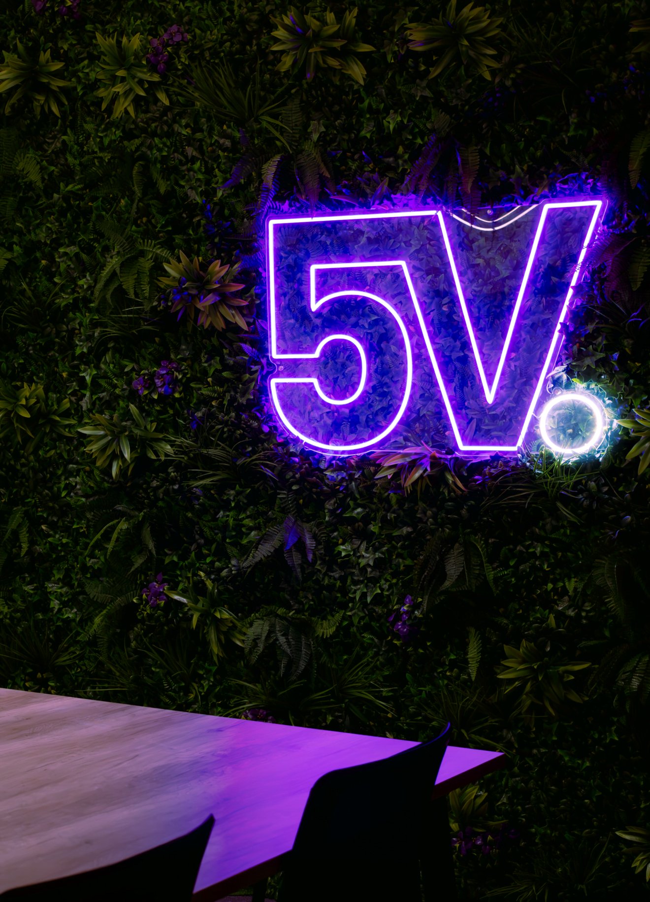 A purple neon sign on a feature wall with plants and flowers on it.