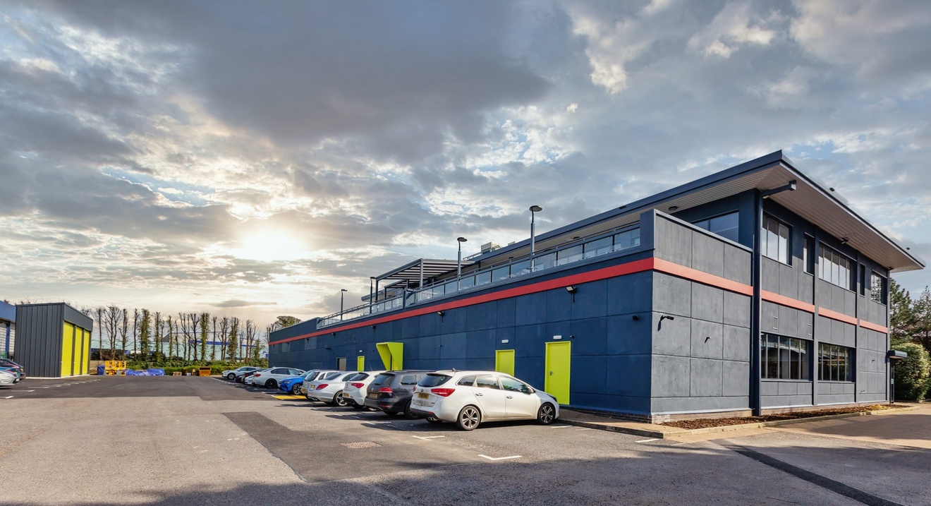 car park with multiple cars parked with cloudy skies and modern squared off grey building with large windows and second floor patio