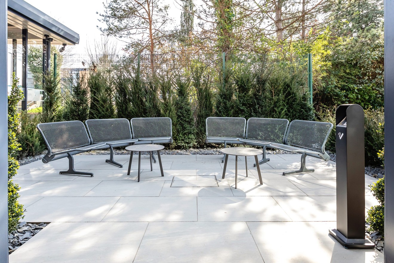 metal chairs and tables outside at a patio with a cigarette bin surrounded by trees and plants