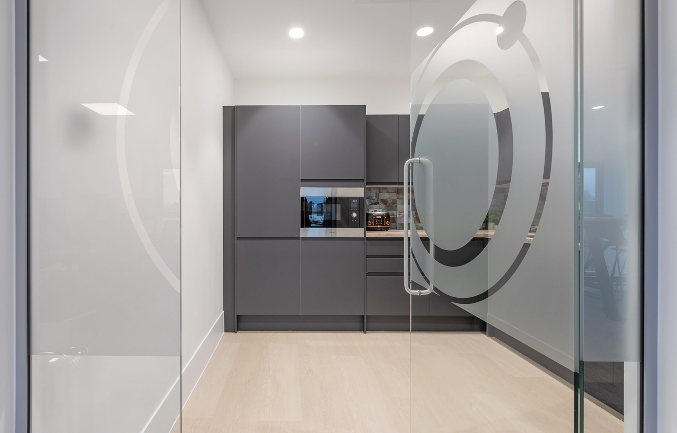 glass door with logo design open leading to a modern minimalist dark grey and white kitchen with cupboards and appliances