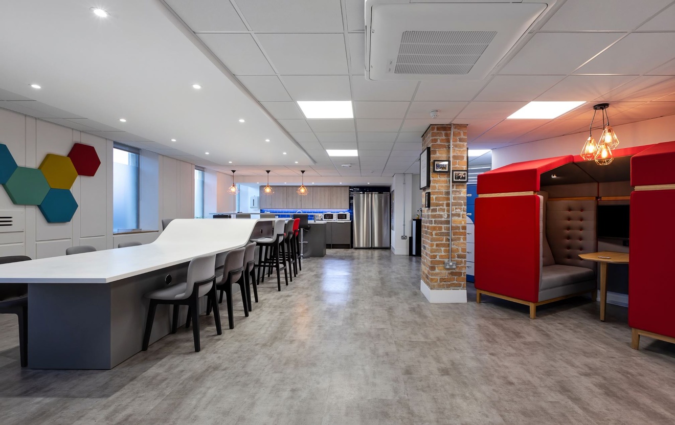 A long table and stools on the left, private working pods on the right, a kitchenette along the back wall and brick support beams in the centre.