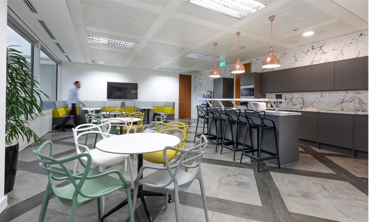 An office kitchen space with a bar and stools, sofa booths and tables with chairs.