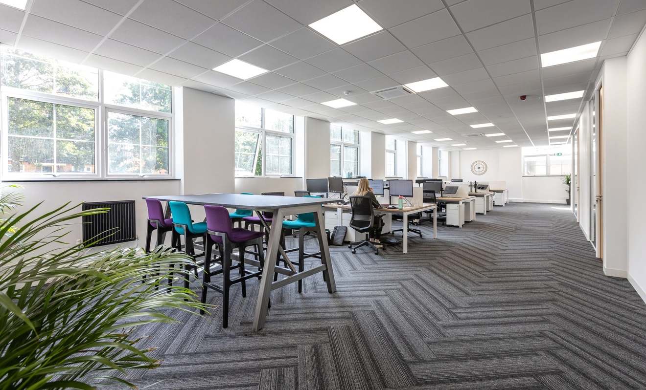 An office space with lines of desks a patterned carpet, white walls and a separate table and stools.