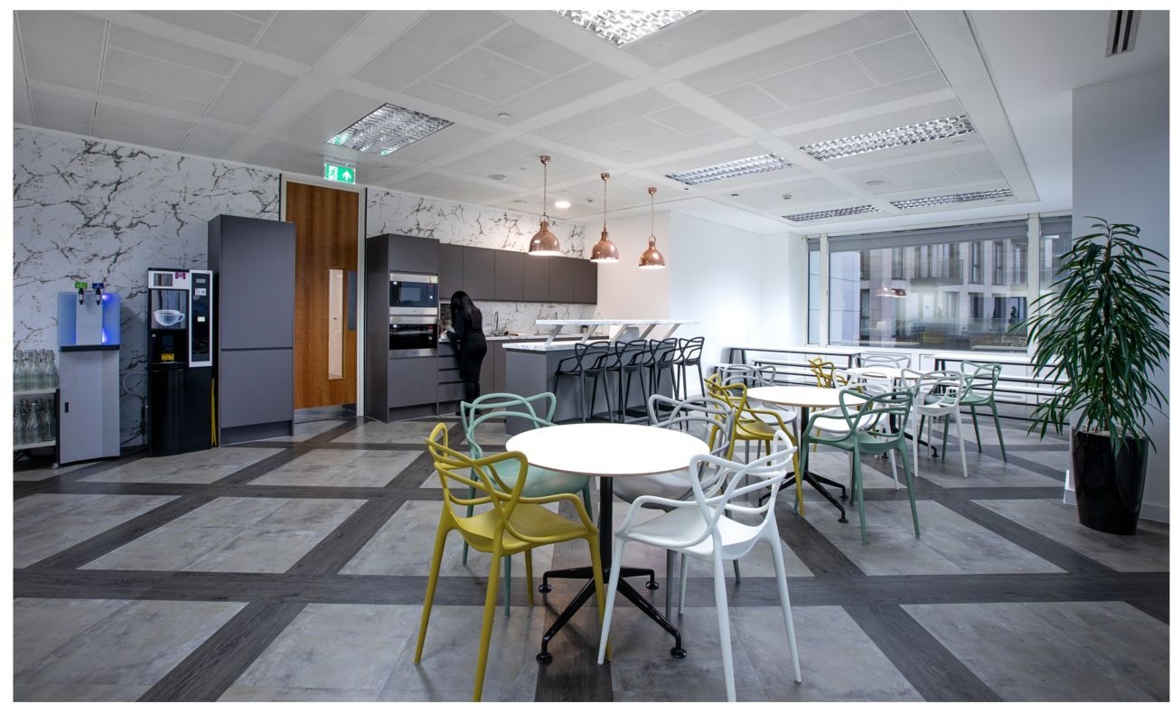 An office kitchen space with a person stood at the work top with a bar, stools and table with chairs in the background.