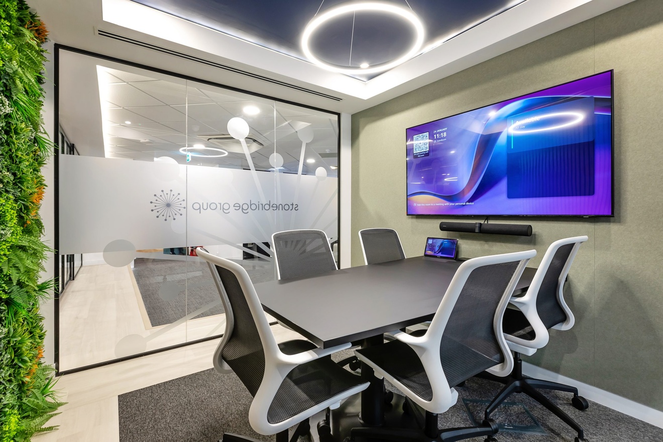 small meeting room with large flat screen tv and soundbar with black table and five swivel chairs with plants on the left wall
