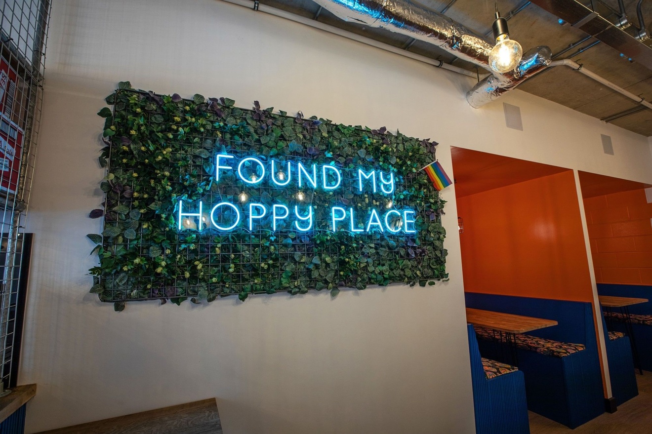 A Hoppy Place neon sign surrounded by green foliage.