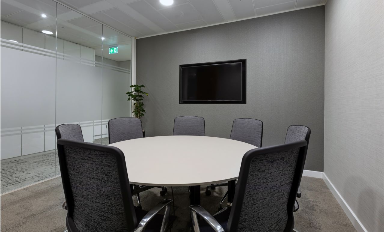 A round boardroom table, surrounded by chairs and a wall-mounted tv.