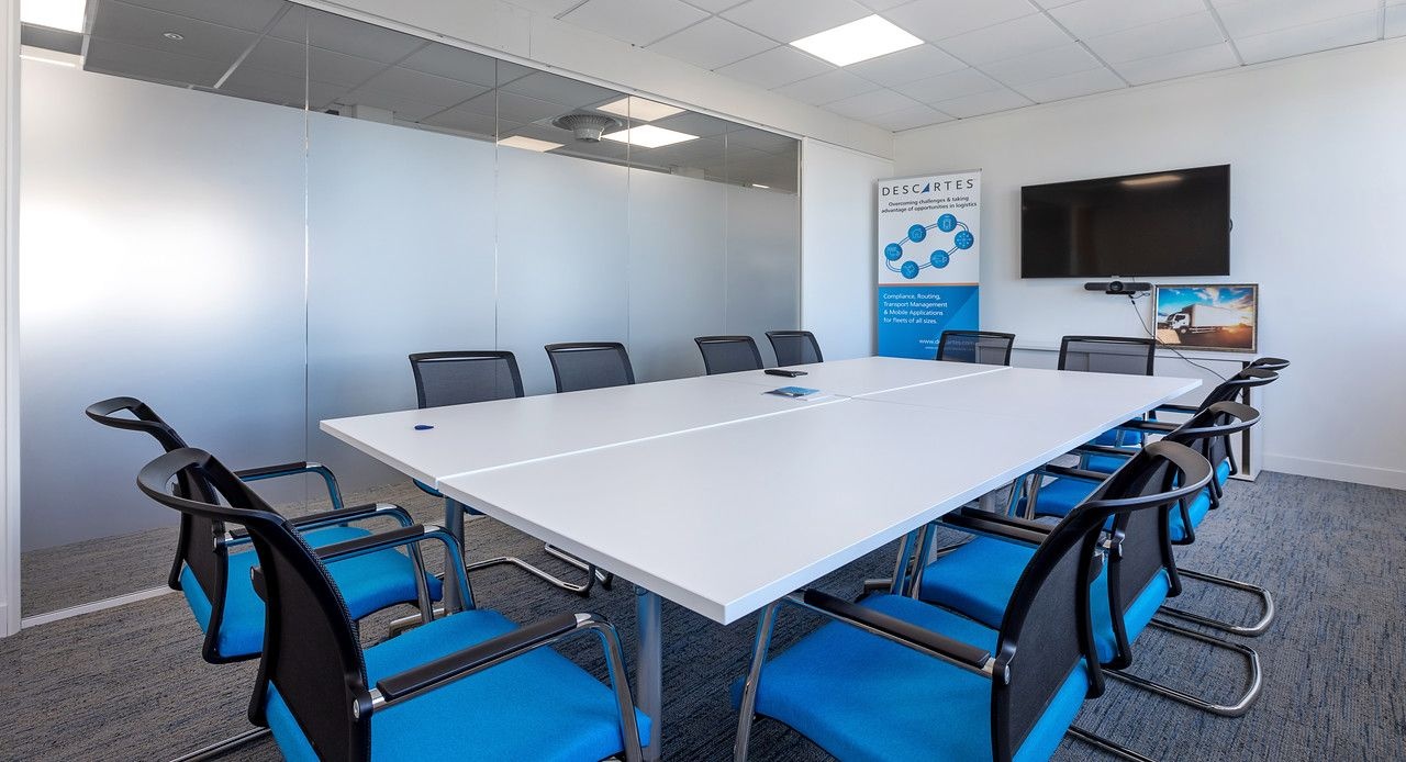 A meeting room with a white board table, surrounded by blue and black chairs and a television on the back wall.